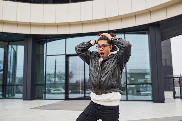 Handsome young man with curly black hair standing and feels shoked on the street against building.