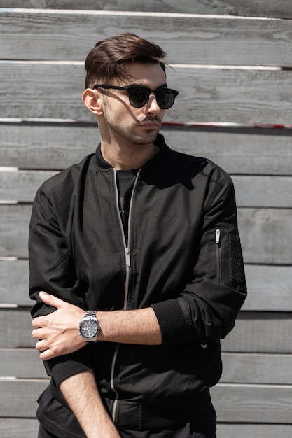 Handsome young man with fashionable hairstyle in trendy black jacket in a stylish dark sunglasses poses near a gray vintage wall in a sunny summer day. Attractive brunette guy outdoors.