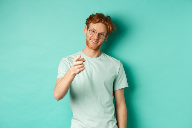 Handsome young man with ginger hair, wearing glasses and t-shirt, pointing finger at camera and smiling, choosing you, making invitation, standing over turquoise background