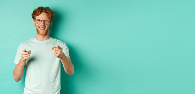 Handsome young man with ginger hair wearing glasses and tshirt pointing finger at camera and smiling