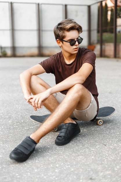 Photo handsome young man with the hairstyle in fashion sunglasses sitting on a skateboard.