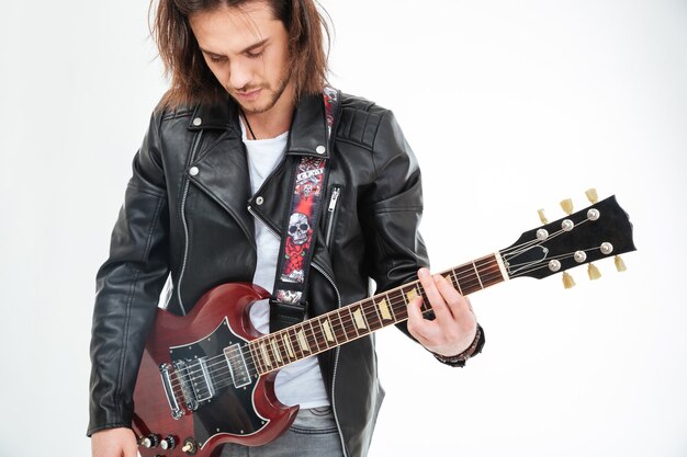 Handsome young man with long hair in black leather jacket playing electric guitar over white background