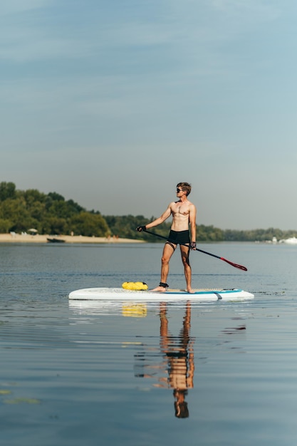 Красивый молодой человек с обнаженным туловищем стоит на доске с веслом в руке и плывет