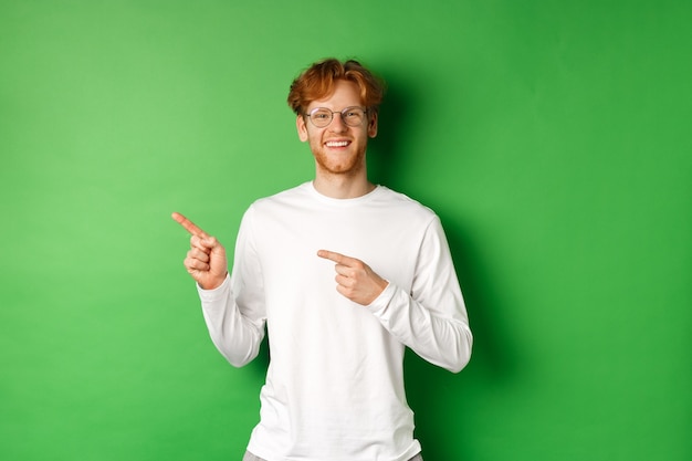 Красивый молодой человек с рыжими волосами и бородой улыбается, указывая пальцами влево на логотип, показывая рекламу, стоя на зеленом фоне.