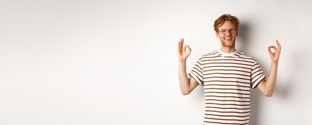 Handsome young man with red messy hair and glasses smiling showing ok signs in approval praising som