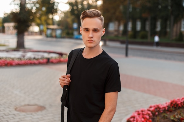 Handsome young man with a stylish hairstyle in a black fashionable t-shirt with a stylish backpack walks around the city