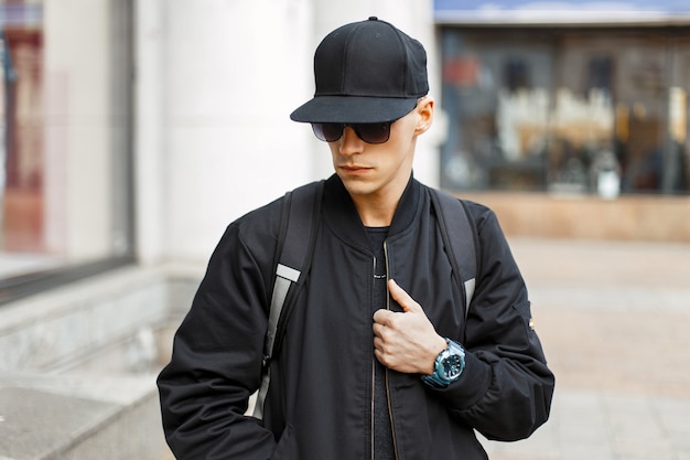 Handsome young man with sunglasses in a black jacket with a backpack in the city