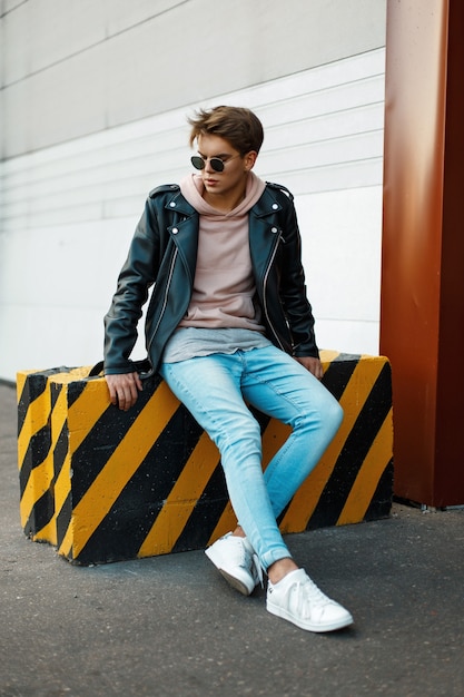 Handsome young man with sunglasses in a black leather jacket, blue jeans and white sneakers sits on a black-and-yellow slab