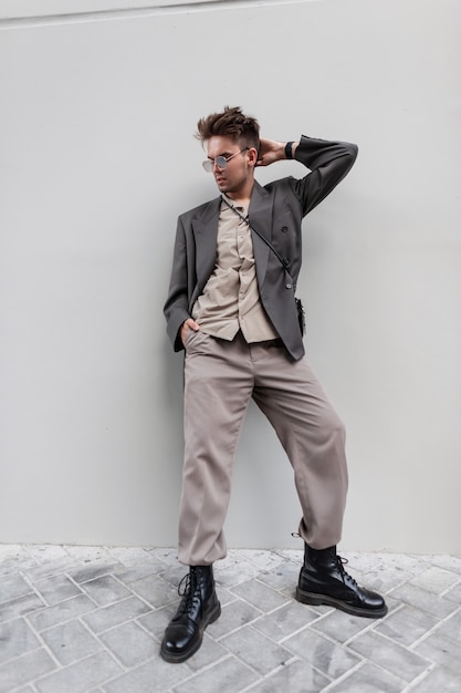 ファッション衣装のサングラスをかけたハンサムな若い男は、通りの灰色の壁の近くにブレザー、シャツ、革の靴を履いています。アーバン男性スタイルのファッション