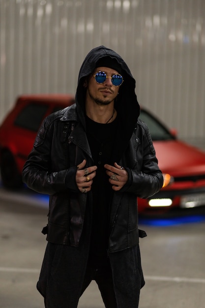Фото Красивый молодой человек в модных синих солнцезащитных очках в стильной кожаной куртке с модной толстовкой возле красной машины на улице ночью