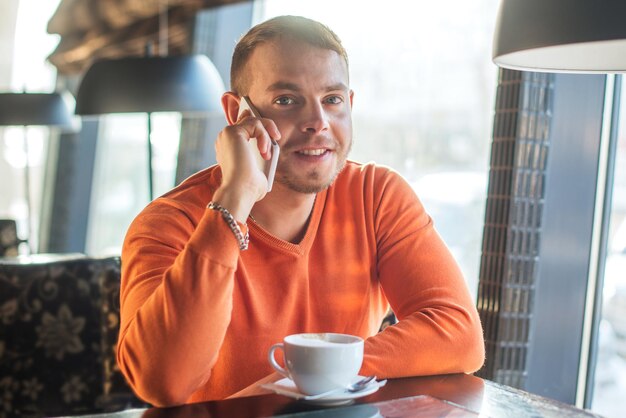 電話で話し、カフェでコーヒーを楽しみながら笑顔で働いているハンサムな若い男はカメラを見てください