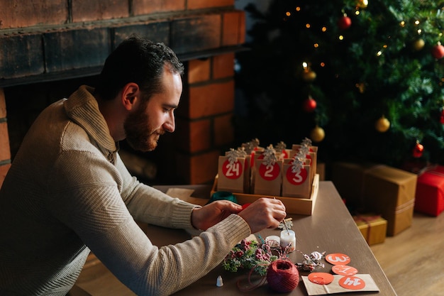 Handsome young man writing number on red bag making paper bags from kraft paper for advent calendar on Christmas Eve sitting by fireplace and tree