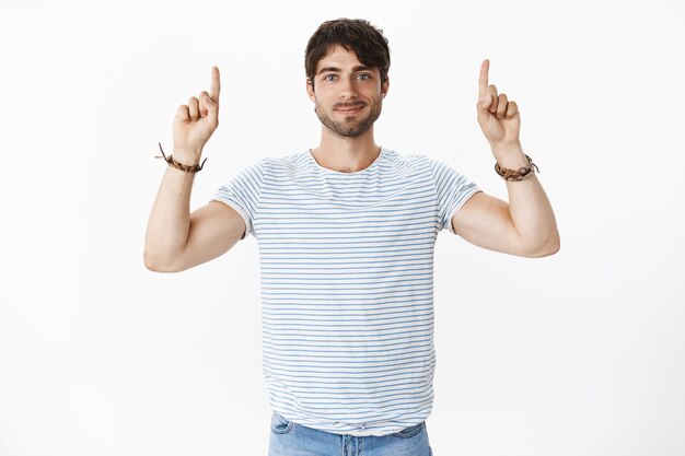 Handsome young, masculine guy with big blue eyes smiling friendly with raised hands pointing up suggesting copy space, checking out cool place for advertising over gray wall