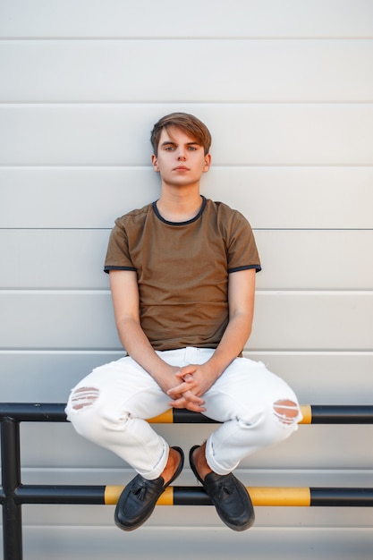 Photo handsome young model man in classic t-shirt with white pants and black shoes sitting near a modern wall