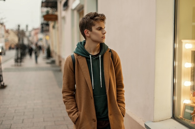 Handsome young model of a man in a trendy coat with a hoodie walks in the city near the shop window