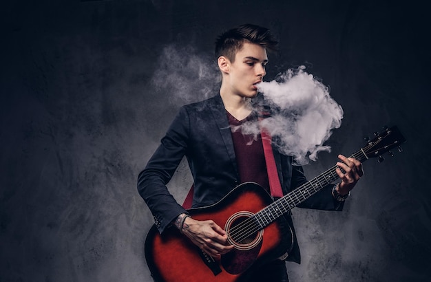 Handsome young musician with stylish hair in elegant clothes exhales smoke while playing acoustic guitar. Isolated on a dark background.