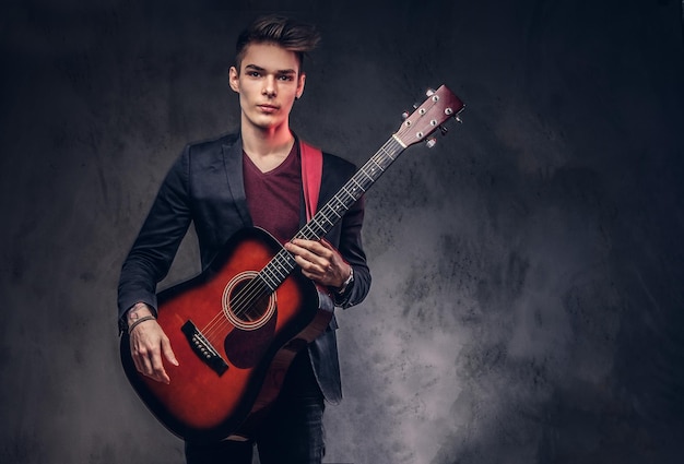 Handsome young musician with stylish hair in elegant clothes with a guitar in his hands playing and posing on a dark background.