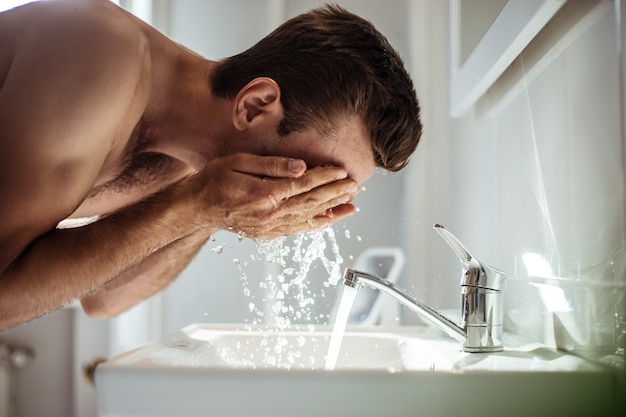 Bel giovane uomo nudo si lava la faccia in bagno a casa.