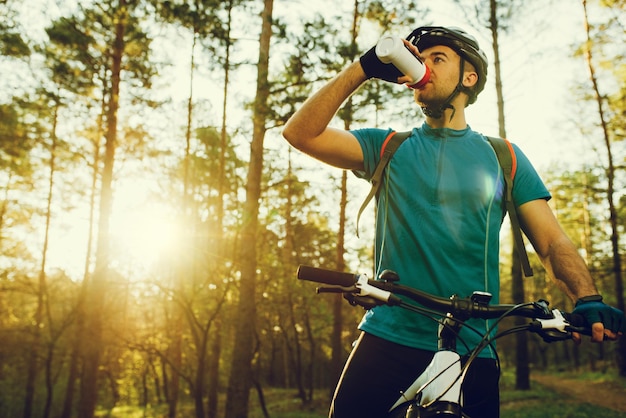 Handsome young professional cyclist dressed in cycling clothing and protective helmet feeling free and happy drinking water from the bottle enjoying bicycle in warm spring Recreation of sportsman