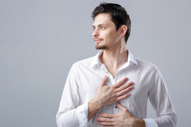 Handsome young proud man in a white shirt holding hand on chest on grey background.