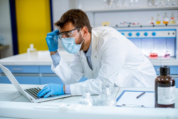 Bel giovane ricercatore in camice bianco e maschera protettiva che lavora utilizzando il computer portatile mentre era seduto in laboratorio