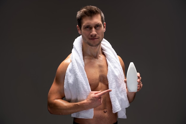 Красивый молодой мускулистый мужчина без рубашки с полотенцем на плечах, указывающий на шампунь в руках, стоя на коричневом фоне. Концепция ухода за мужчинами
