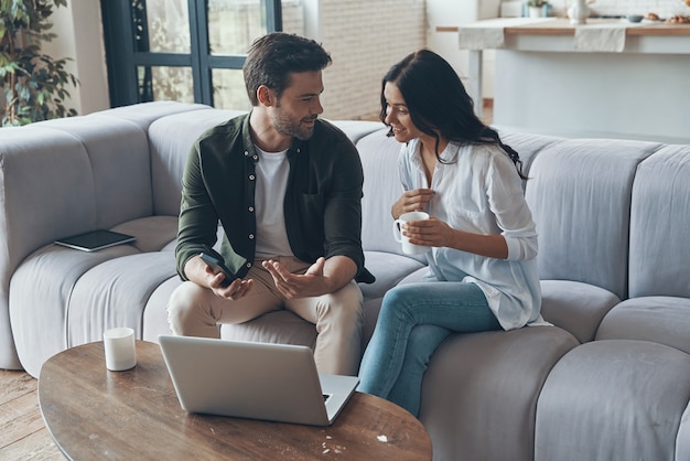 Handsome young showing his girlfriend smart phone and smiling while sitting on the sofa