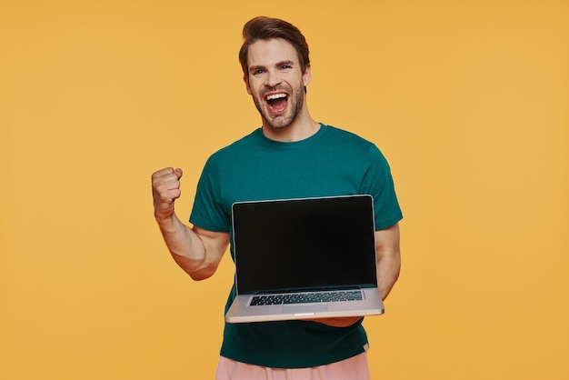 Handsome young smiling man in casual clothing carrying laptop and gesturing while standing against yellow wall