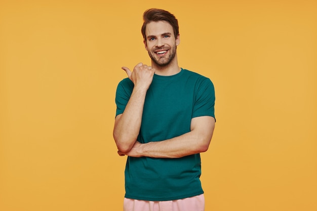 Handsome young smiling man in casual clothing  and pointing away while standing against yellow wall