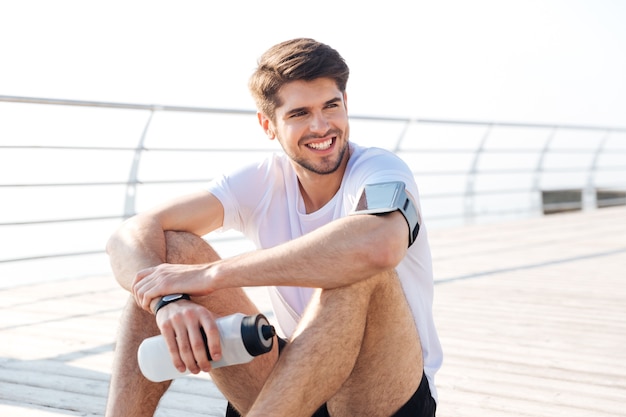 Handsome young smiling sportsman sitting and holding water bottle outdoors