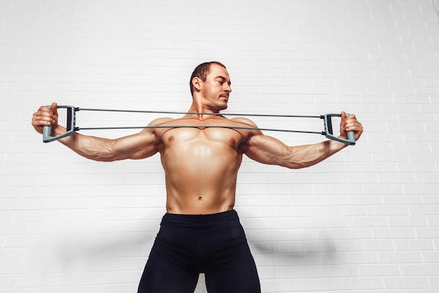 Handsome young sportsman exercising using expander while standing against white background