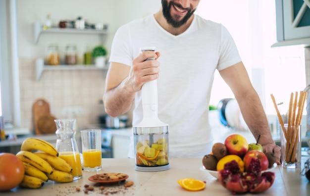 Bel giovane uomo sorridente sportivo in cucina sta preparando un frullato