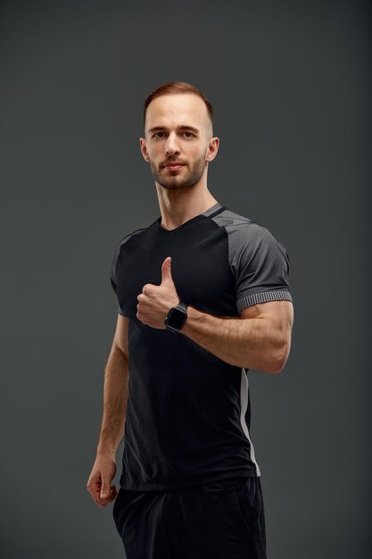 Фото Красивый молодой сильный мужчина модель с мускулистым телом в черном топе показывает большой палец вверх на сером