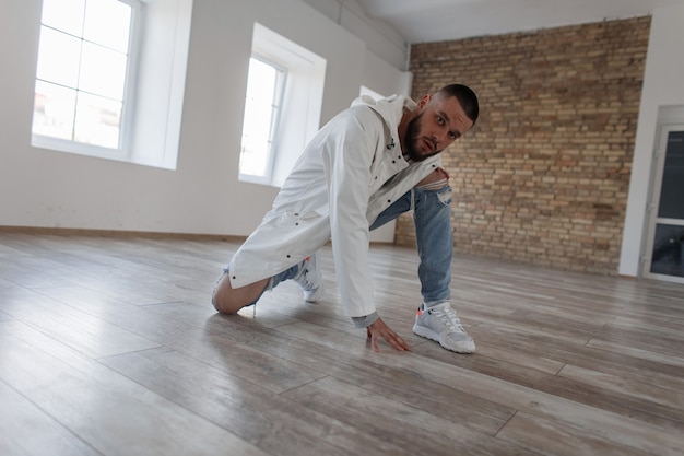 Uomo bello giovane hipster alla moda con la barba in una giacca alla moda e jeans strappati con scarpe da ginnastica bianche seduto e in posa in uno studio di luce diurna