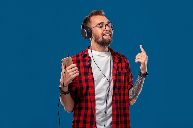 Handsome young stylish man in headphones holding mp player and dancing while standing against blue b...