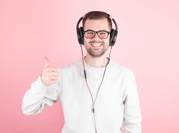 Handsome young stylish man in headphones listening to good music, showing super sign, standing on pink