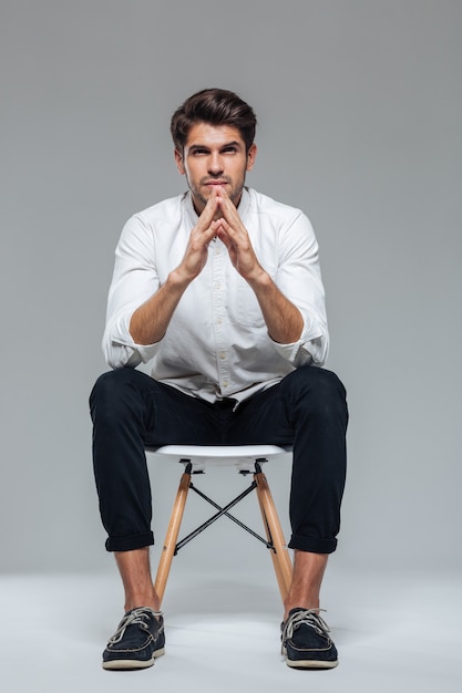 Foto bel giovane uomo premuroso in camicia bianca seduto sulla sedia e guardando davanti isolato su un muro grigio