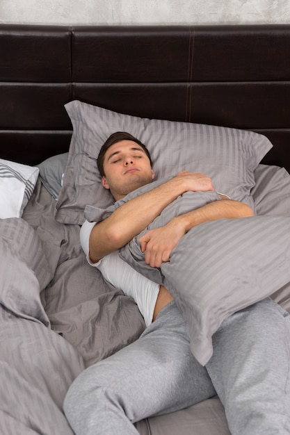 Handsome young tired man in pajama sleeping alone without blanket and hugging a pillow in stylish bed in grey colors and near bedside table with candles in a bedroom in loft style