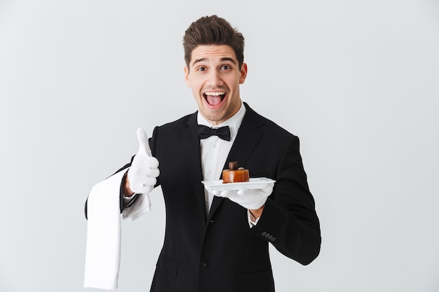 Handsome young waiter in tuxedo with bowtie holding plate with cake isolated over white wall, thumbs p