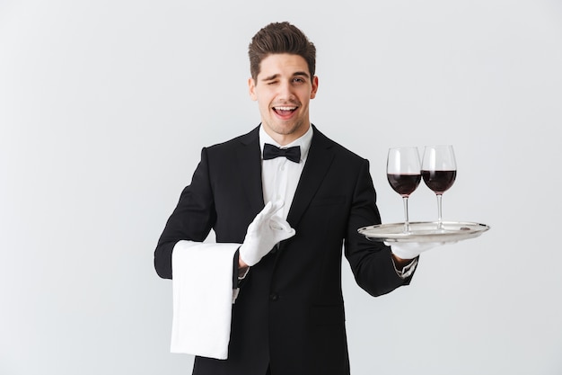 Handsome young waiter wearing tuxedo presenting a tray with two glasses of red wine isolated over gray wall