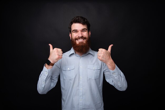 A handsomea bearded guy with toothy smile, showing thumbs up while looking at camera, standing over dark isolated background