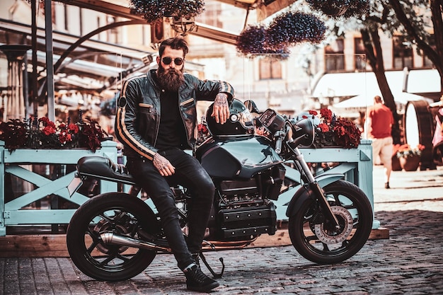 Красивый серьезный байкер с бородой и татуировками сидит на своем мотоцикле в оживленном центре города.
