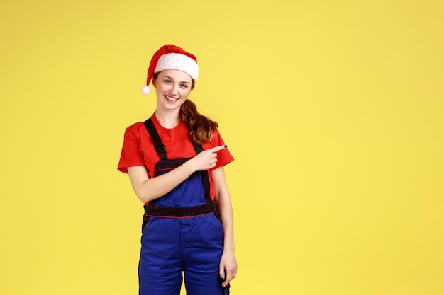 Handy woman looks at camera with smile pointing finger aside showing copy space for advertisement wearing blue overalls and santa claus hat Indoor studio shot isolated on yellow background