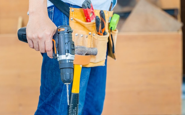 Фото Разнорабочий в поясе с инструментами и держащий дрель в руках плотник, работающий с оборудованием в столярной мастерской