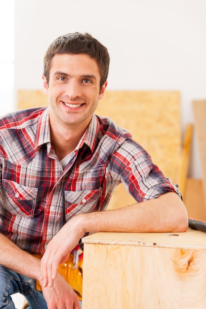 Tuttofare che si rilassa dopo il lavoro. bel giovane tuttofare che guarda la macchina fotografica e sorride mentre è seduto in officina e si appoggia al ponte di legno