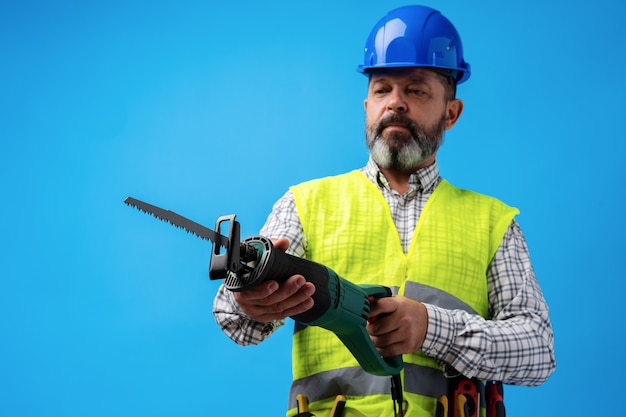 Handyman in uniform holding saw against blue background