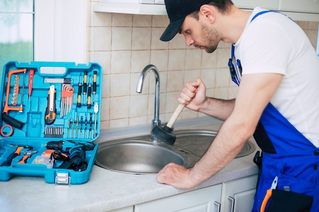 Handyman in uniform is cleaning a clogged kitchen sink with the help of plunger