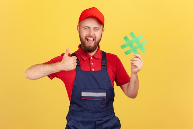 Handyman wearing blue overalls standing holding green hashtag and showing thumb up