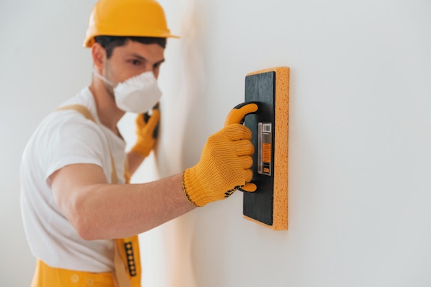 Handyman in yellow uniform and protective mask polishing wall indoors House renovation conception