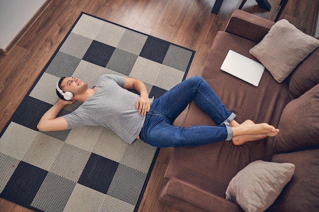 Hanfsome young male staying alone on floor with legs on couch while enjoying time in headphones